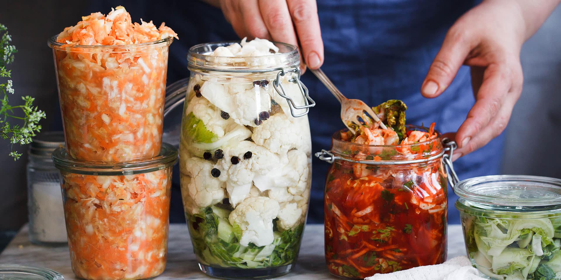 Hands place fermented kimchi into a jar, which sits next to jars of other pickled and canned vegetables.